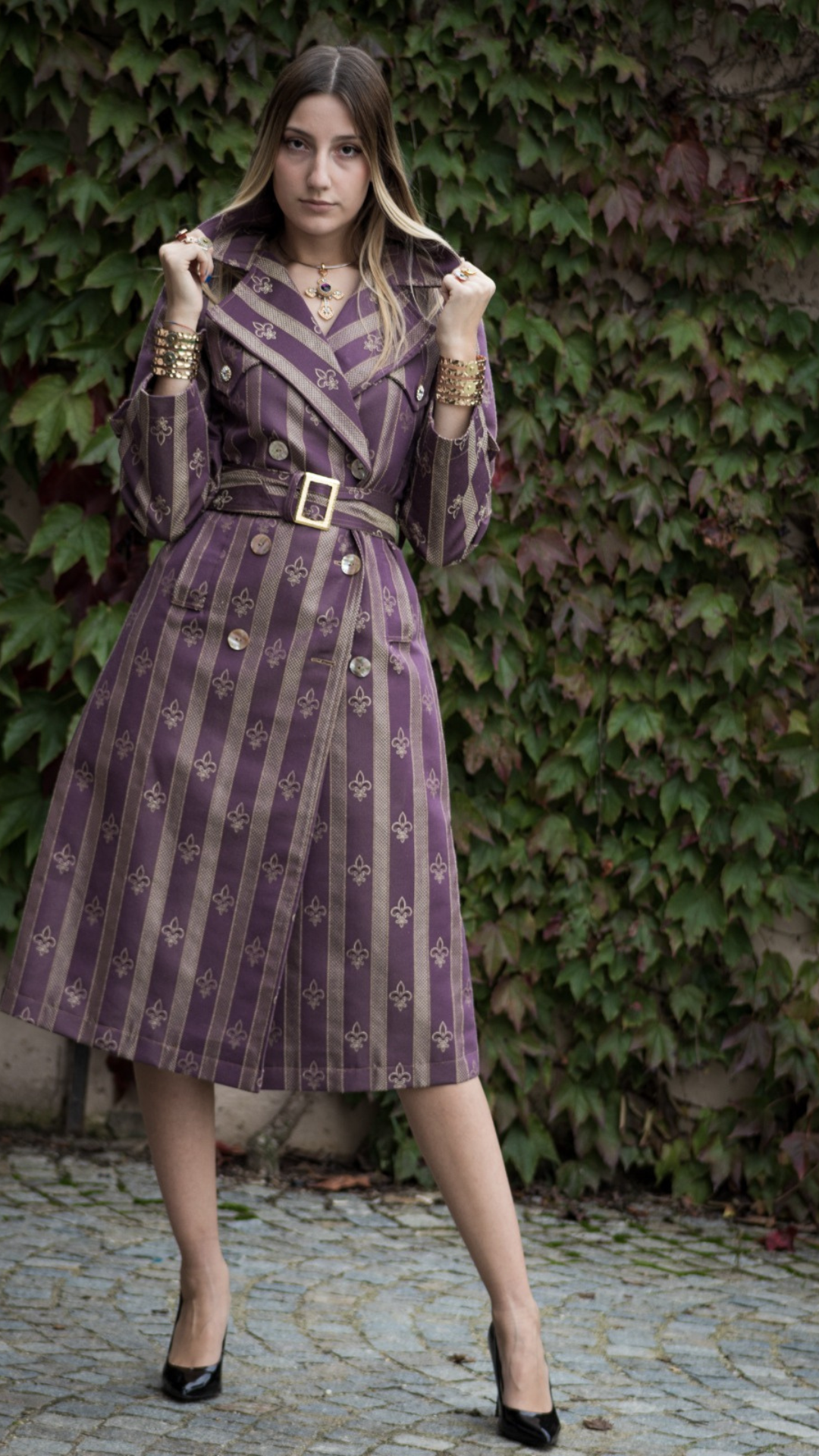 Donna con trench viola a motivi dorati, accessoriata con bracciali dorati e collana con croce colorata, posa con grinta su sfondo di edera mettendo in evidenza i Gioielli Calamita di Claudia De Rosa Jewelry