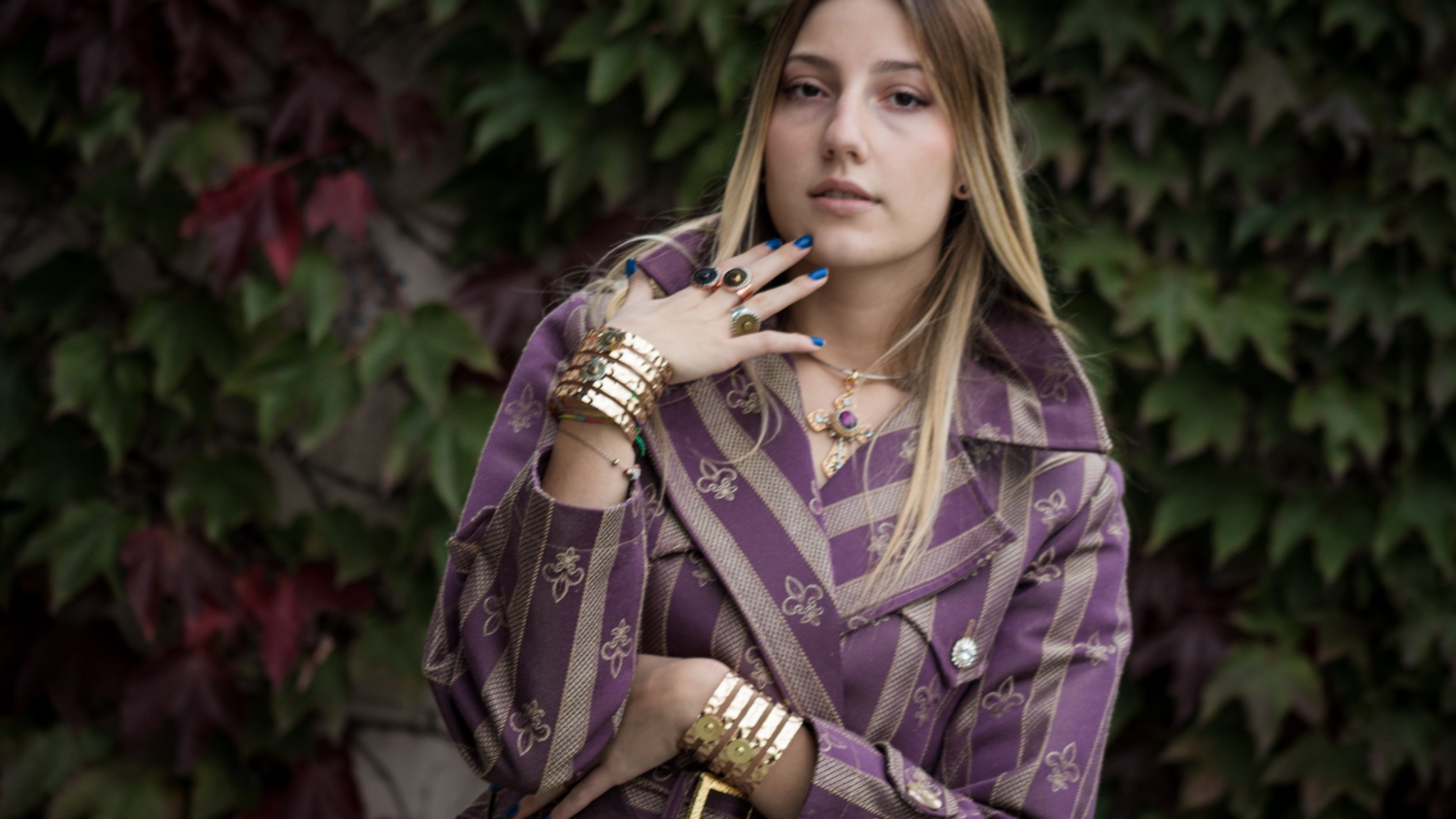 Donna con trench viola a motivi dorati, accessoriata con bracciali dorati e collana con croce colorata, posa con grinta su sfondo di edera.